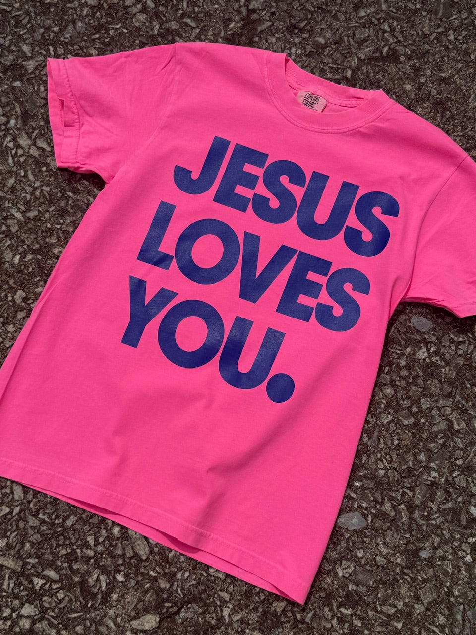 Hot Pink short sleeve t-shirt with Large 'Jesus Loves You' text printed in navy blue. Shirt is laying on floor.