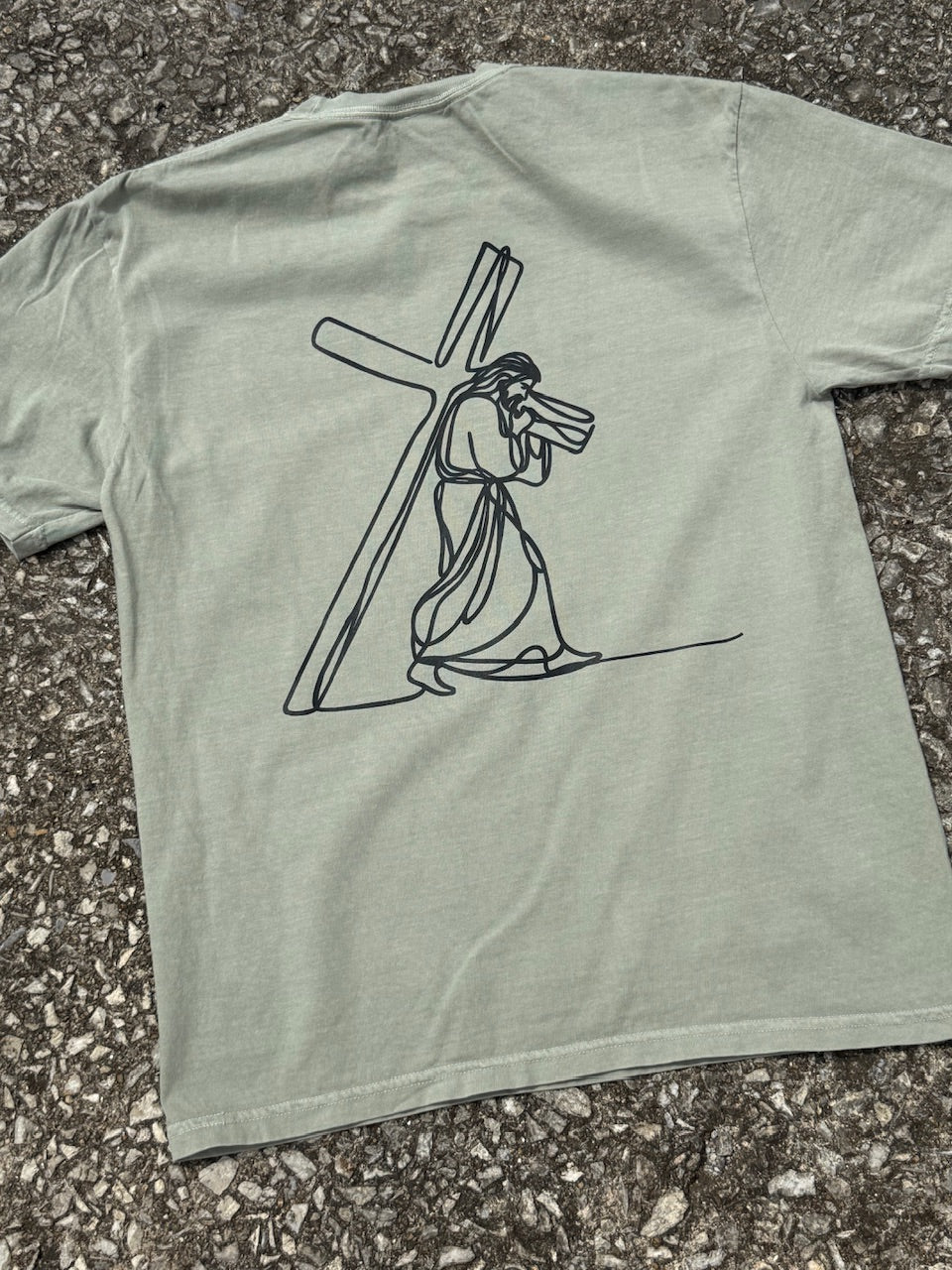 The backside of a Light green short sleeve T-shirt with graphic of Jesus carrying a cross printed in black. Shirt is laying on the ground.