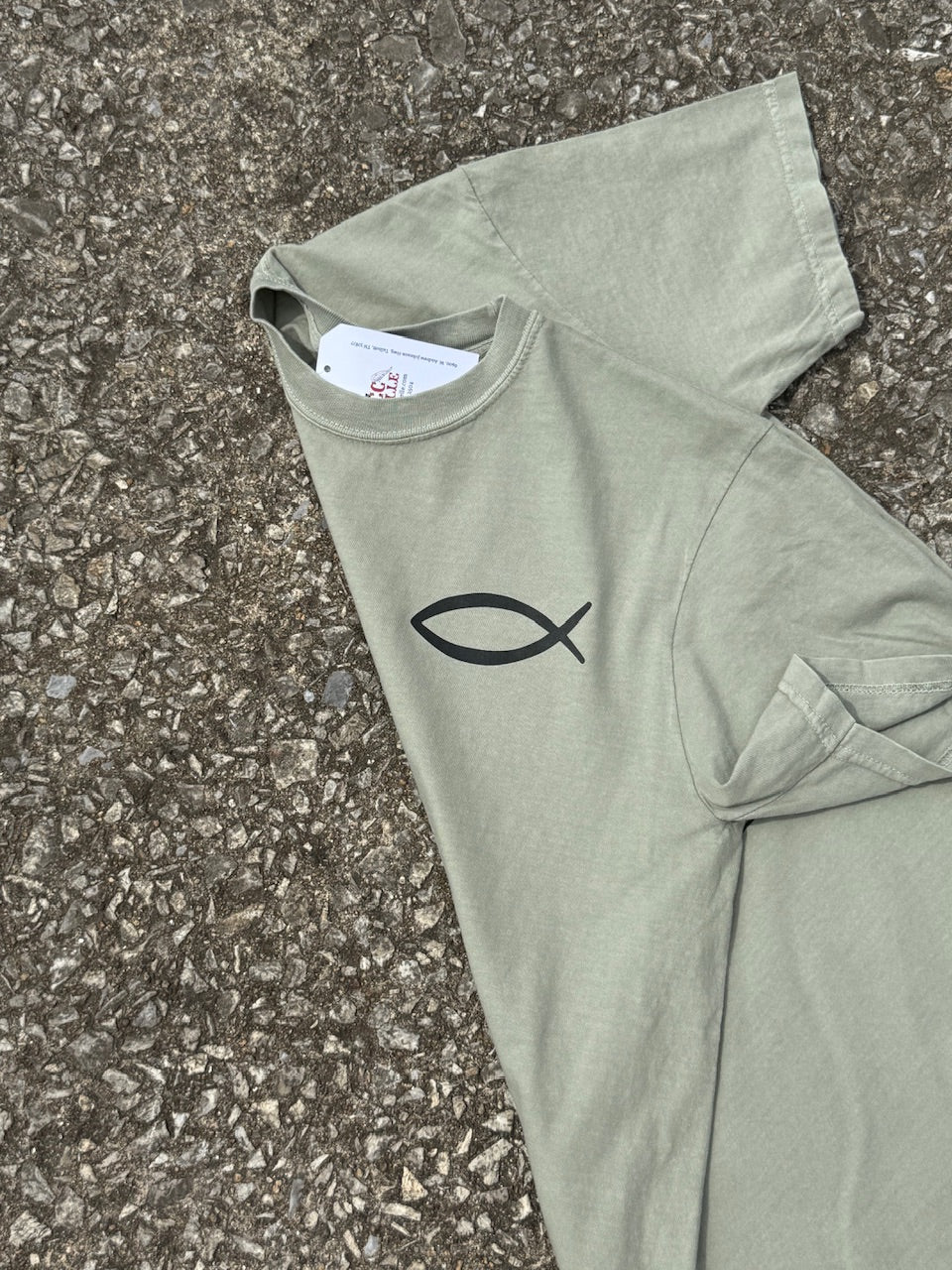 The front of a light green short sleeve T-shirt that has a small christian fish graphic printed in black on the top left chest area. The shirt is folded and laying on the ground.