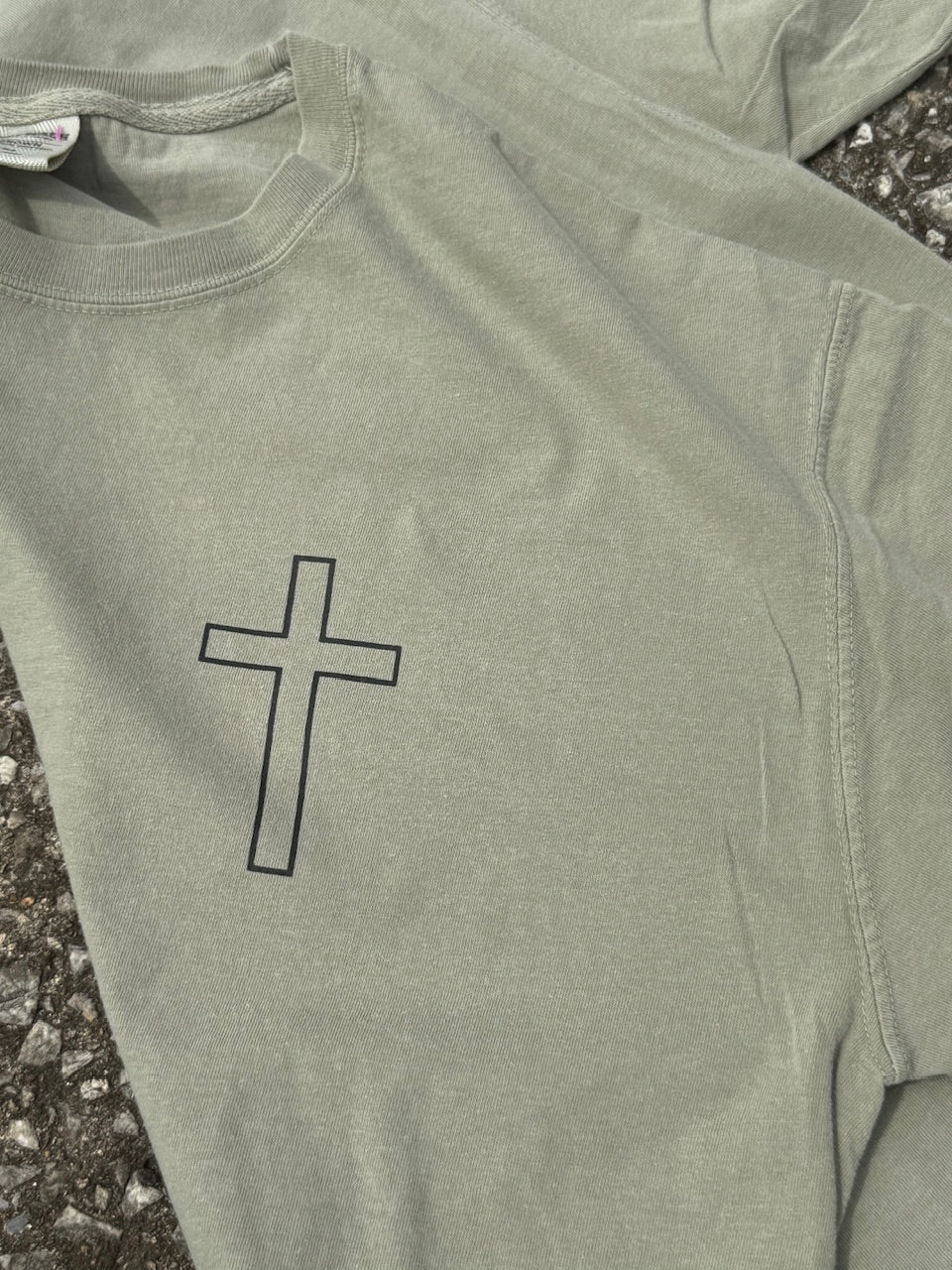 Close up of front of light green short sleeve T-shirt that has a small black cross outline graphic printed in the top left chest area.
