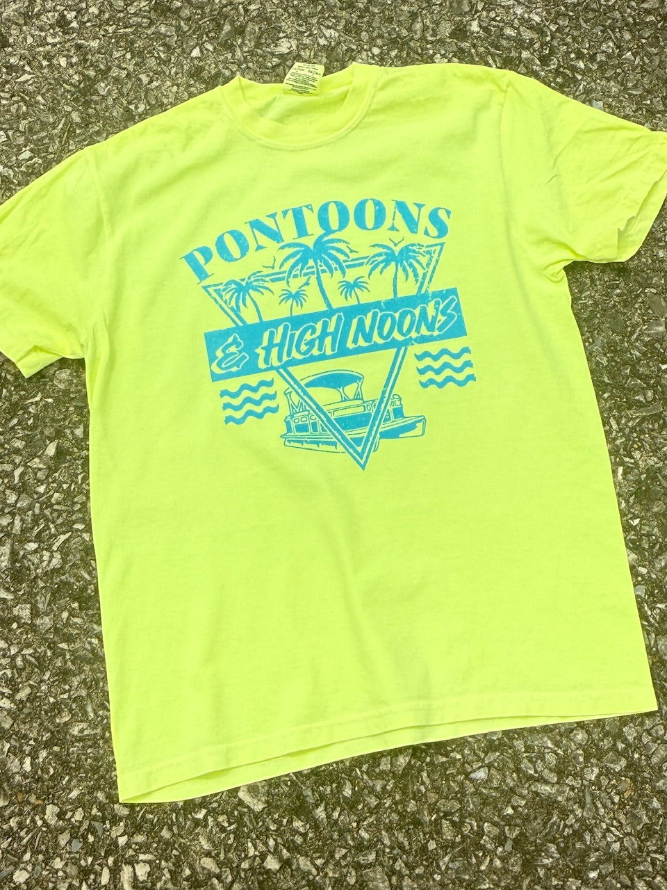 Front of neon yellow short sleeve T-shirt that has the text "Pontoons & High Noons" with a pontoon boat and palm trees in an 80s look printed in teal blue. The shirt is laying on the ground.