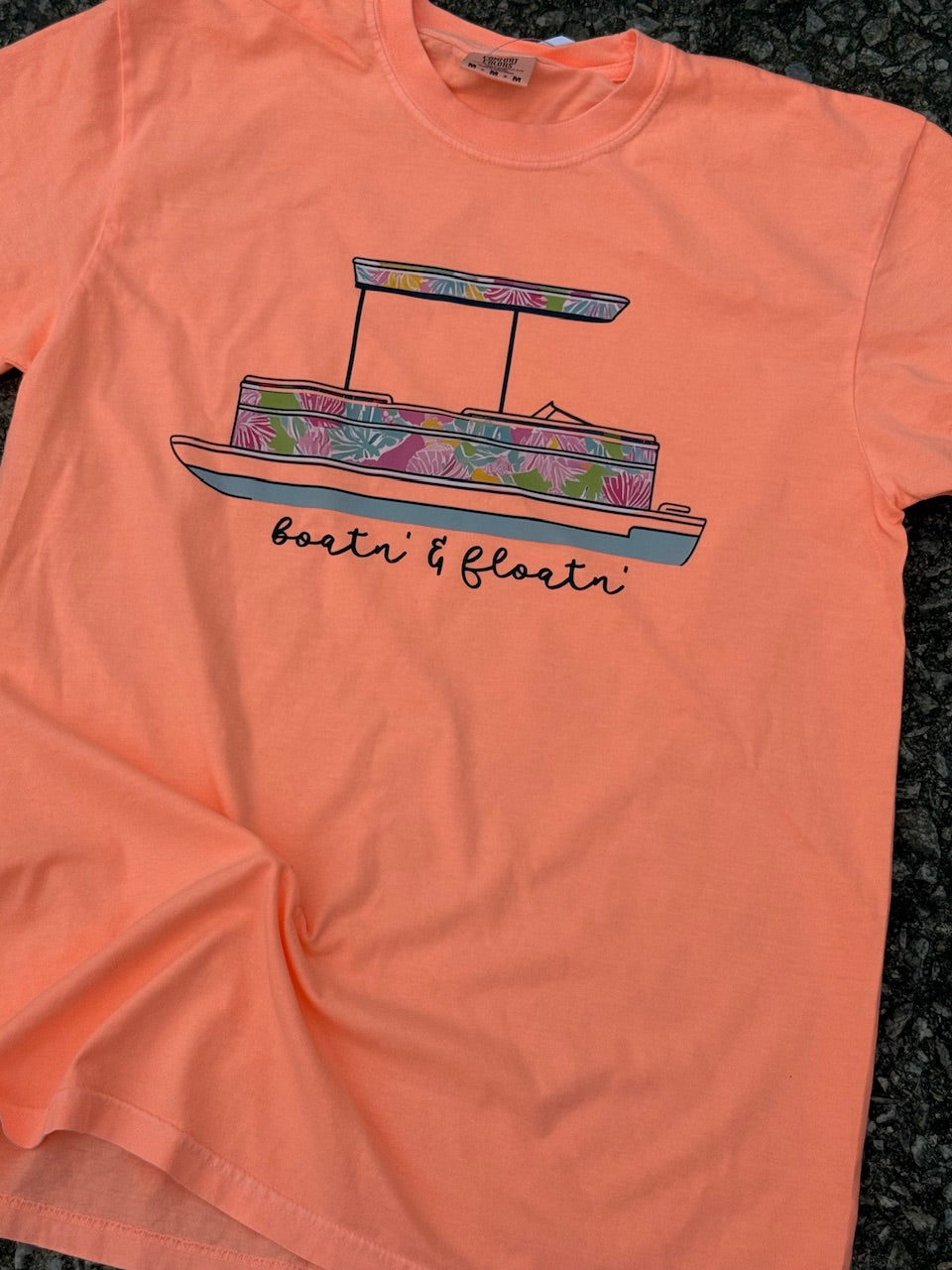 Front of Orange short sleeve t-shirt with a summer floral pontoon boat graphic and 'boatin' & floatin'' text. The shirt is laying on the ground.