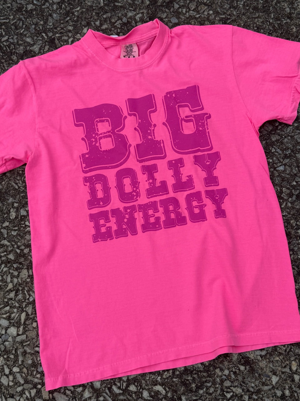 Front of Hot pink short sleeve T-shirt with "Big dolly energy" in a western font printed in dark pink. The shirt is laying on the floor.