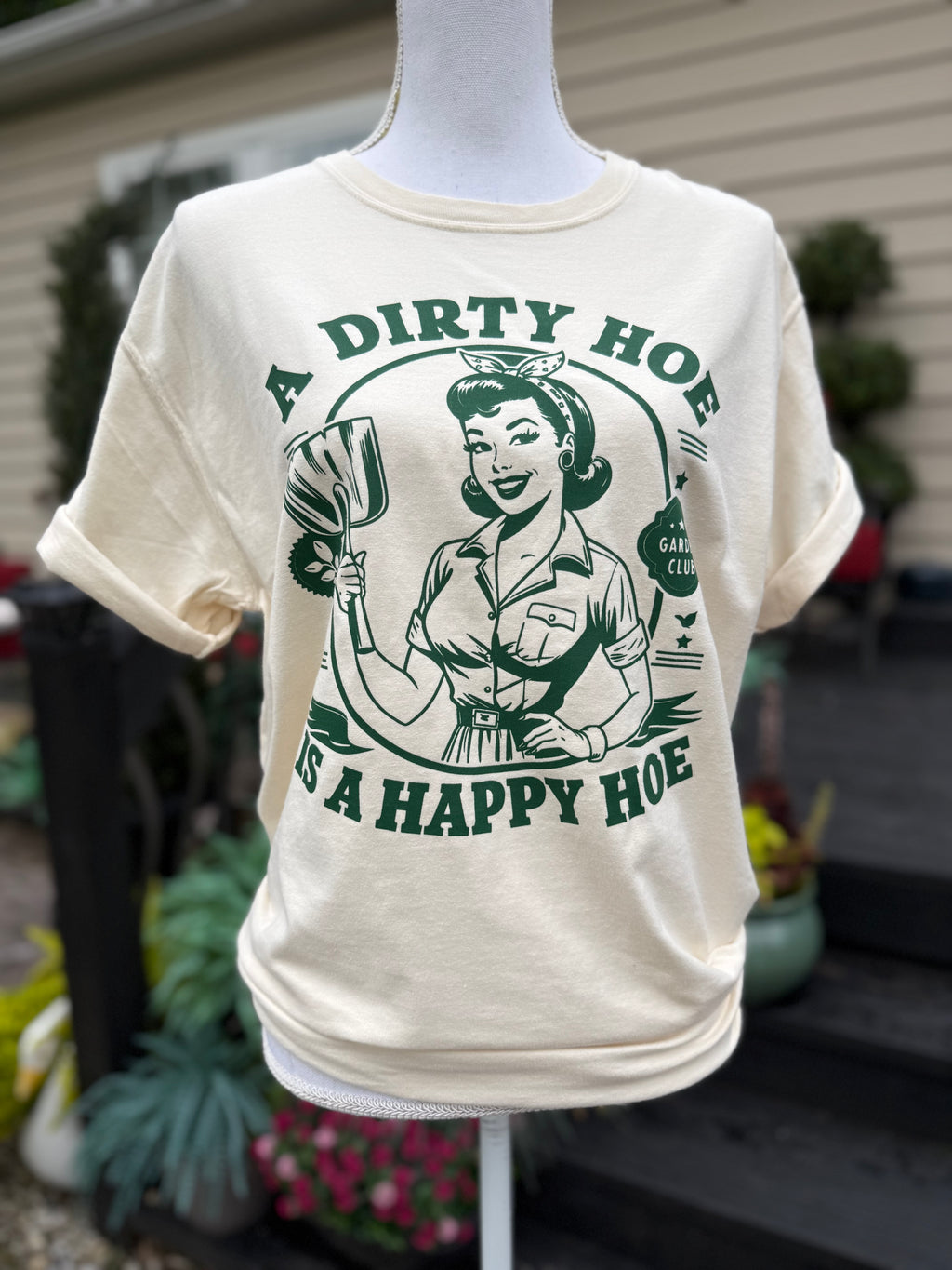 Cream T-shirt with a vintage lady that is holding a trowel with the text "A dirty hoe" above her and " is a happy hoe" below her is printed on the shirt in dark green. A mannequin is wearing the shirt standing in front of blurry plants.