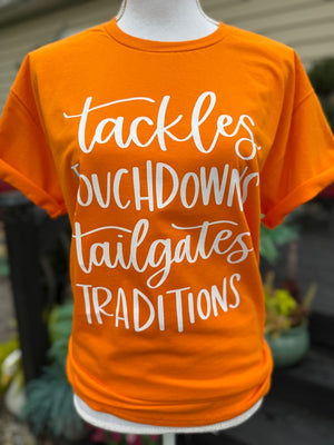 Orange t-shirt with white text that says "Tackles, Touchdowns, Tailgates, Traditions." on a mannequin against a blurred outdoor background