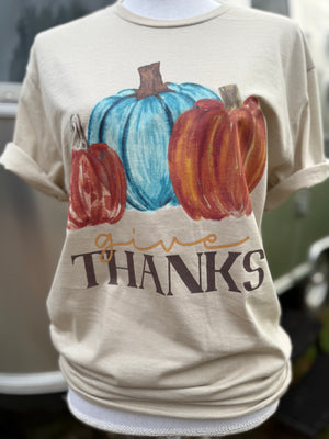 Front of light beige short sleeve T-Shirt with graphic of two orange pumpkins with a teal pumpkin in the middle with the text "give" printed in orange and "THANKS" printed in brown.