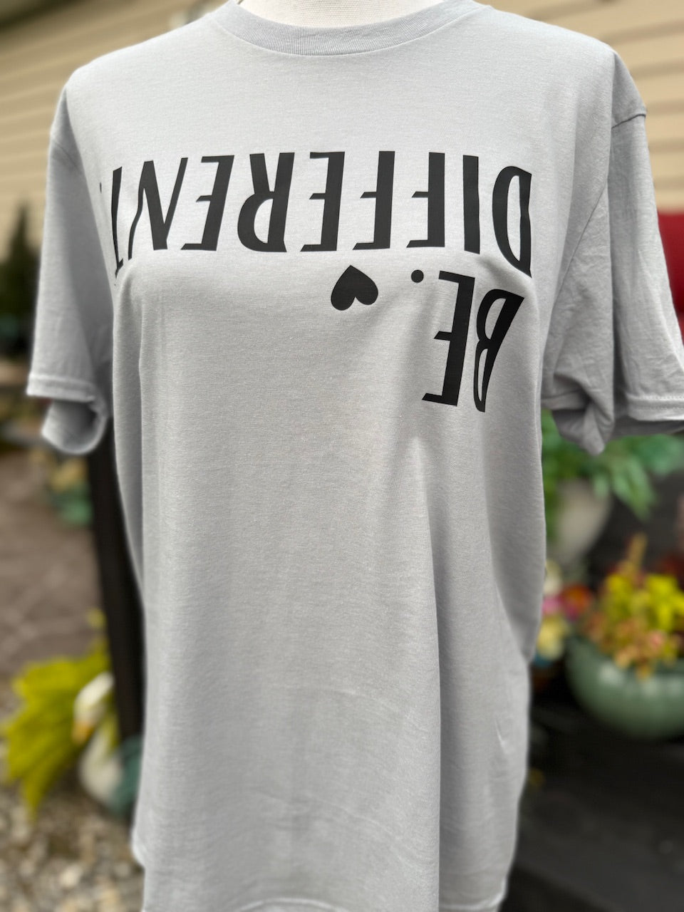 Light grey T-shirt with the text "Be different" with a small heart graphic located next to "Be" is printed on the shirt upside down in black ink. A mannequin is wearing this shirt in front of a blurry background.