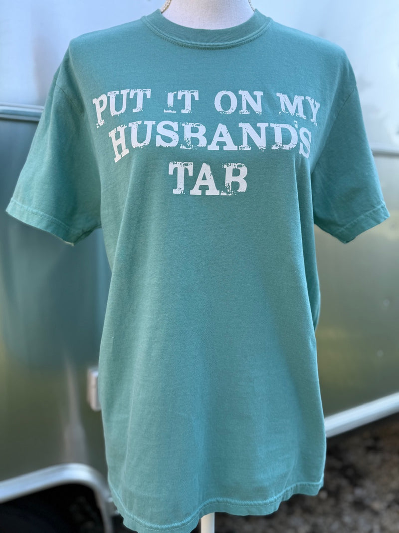 Green T-shirt with the text "put it on my husband's tab." in a distressed font  is printed on the shirt in white ink. A mannequin is wearing this shirt standing in front of a camper.