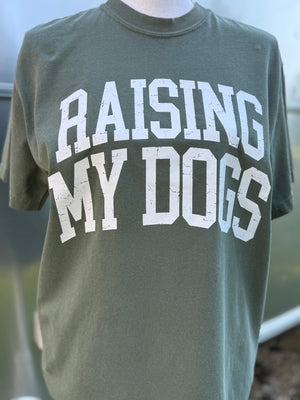 Green t-shirt with 'RAISING MY DOGS' text in white is printed on shirt. A mannequin is wearing this shirt in front of a blurry background.