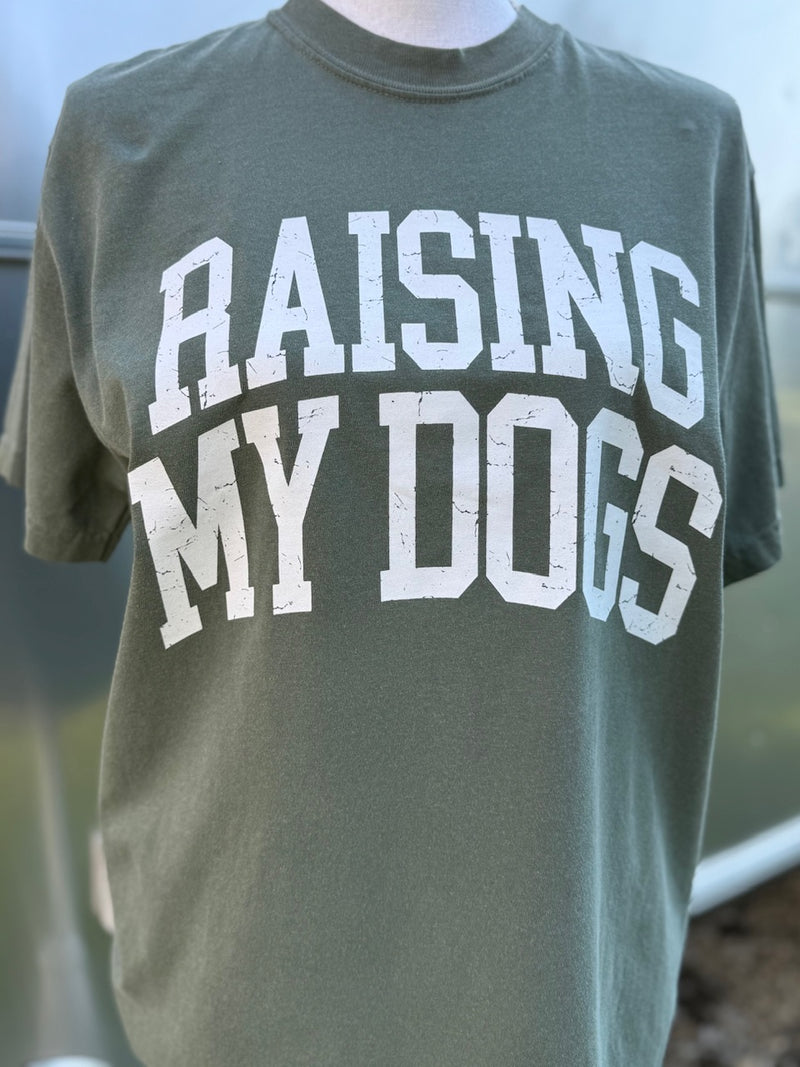Green t-shirt with 'RAISING MY DOGS' text in white is printed on shirt. A mannequin is wearing this shirt in front of a blurry background.