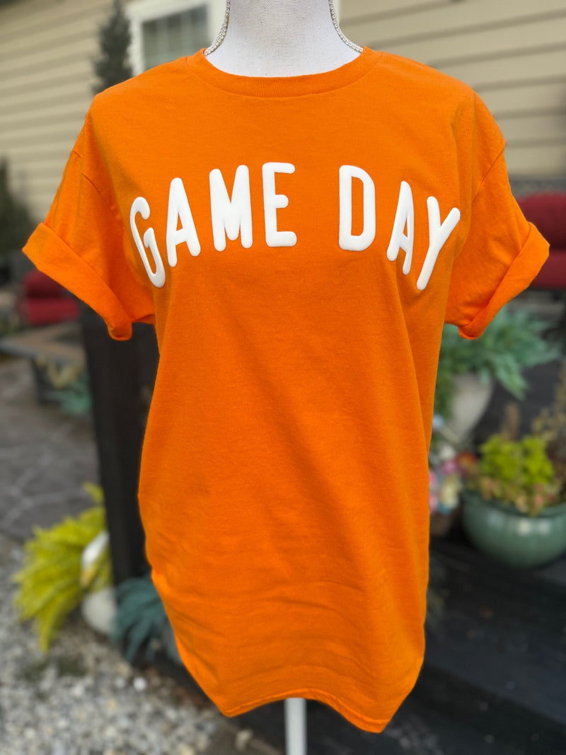 Front of orange short sleeve t-shirt that has the sleeves rolled up with "Game Day" printed in white puffy ink on it. The shirt is on a mannequin in front of a blurry background.
