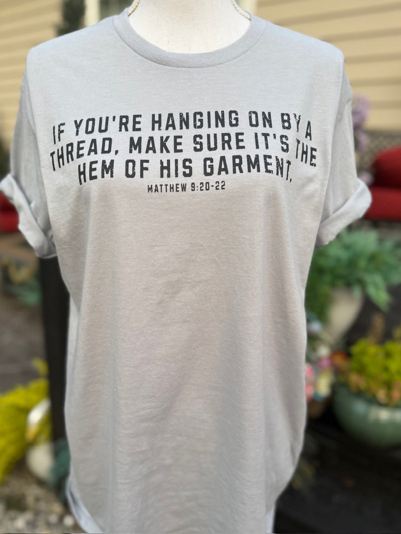 Front of light gray short sleeve t-shirt with the text "If you're hanging on by a thread, make sure it's the hem of his garment. Matthew 9:20-22" is printed in black. The shirt is on a mannequin in front of a blurry background.
