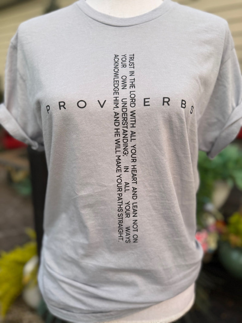 Front of grey short sleeve t-shirt with "Proverbs" printed horizontally and " Trust in the lord with all your heart and lean not on your own understanding , in all your ways acknowledge him, and he wil make your paths straight." printed vertically so that the two texts make a cross is printed in black on the shirt. A mannequin is wearing this shirt in front of a blurry background.