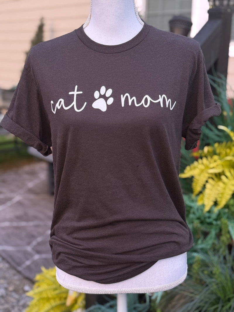 Front of brown short sleeve t-shirt with the text "cat" then a graphic of a paw print then the text "mom" printed in white. The shirt is on a mannequin in front of a blurry background.