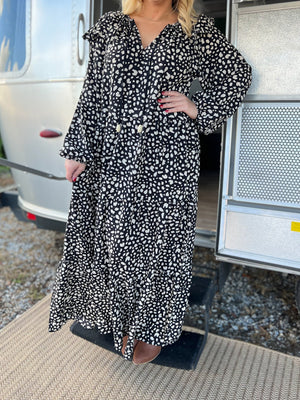 Model is wearing a black dot maxi dress that ties at neck and has ruffles on shoulders